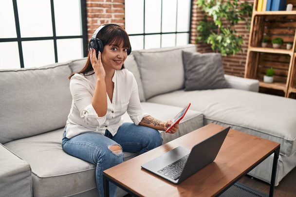 Young beautiful hispanic woman using touchpad and headphones sitting on sofa at home - Foto, Imagen