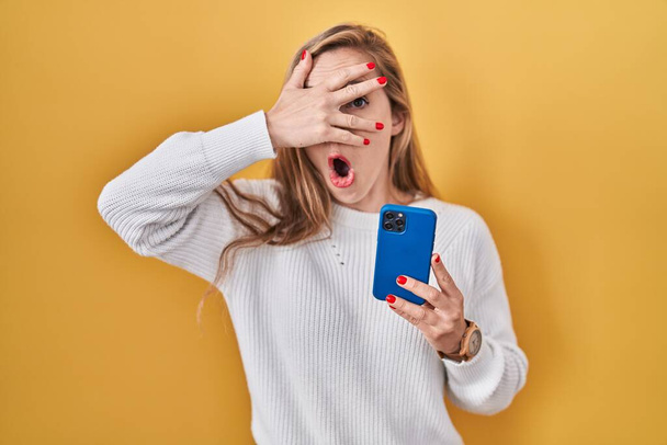 Young blonde woman using smartphone typing message peeking in shock covering face and eyes with hand, looking through fingers with embarrassed expression.  - Fotoğraf, Görsel