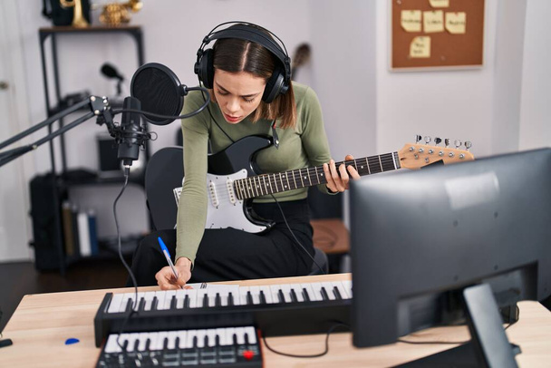 Jonge mooie Spaanse vrouw muzikant componeren lied spelen elektrische gitaar in muziekstudio - Foto, afbeelding