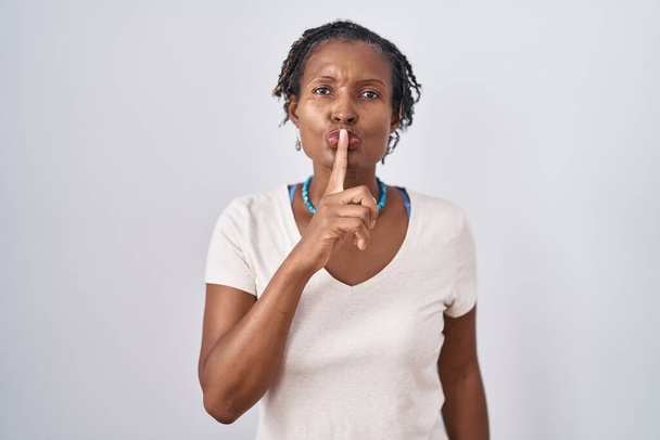唇に指で静かにするように求めて白い背景の上に立っている恐ろしいロックを持つアフリカの女性。沈黙と秘密の概念.  - 写真・画像