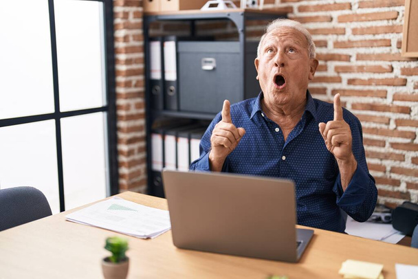 Starší muž s šedivými vlasy pracující pomocí notebooku v kanceláři užaslý a překvapený vzhlíží a ukazuje prsty a zvednutými pažemi.  - Fotografie, Obrázek