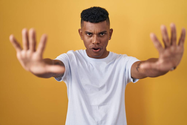 Junger hispanischer Mann steht vor gelbem Hintergrund und macht Stop-Geste mit Handflächen, wütendem und frustriertem Ausdruck  - Foto, Bild