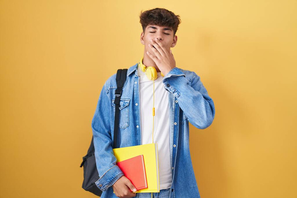 Der hispanische Teenager trägt einen Studentenrucksack und hält gelangweilte Bücher in der Hand, die den Mund mit der Hand gähnend müde bedecken. Ruhelos und schläfrig.  - Foto, Bild