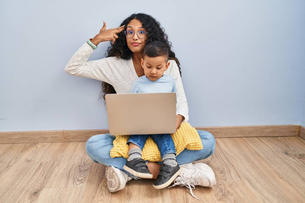 Young hispanic mother and kid using computer laptop sitting on the floor shooting and killing oneself pointing hand and fingers to head like gun, suicide gesture.  - Photo, Image