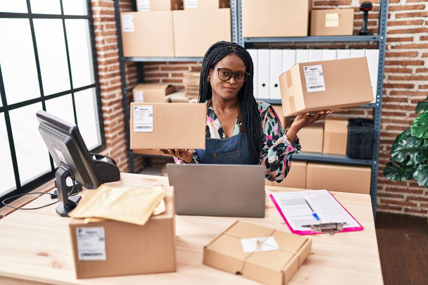 Femme africaine avec des tresses travaillant dans une petite entreprise e-commerce tenant des paquets souriant regardant vers le côté et regardant loin en pensant.  - Photo, image