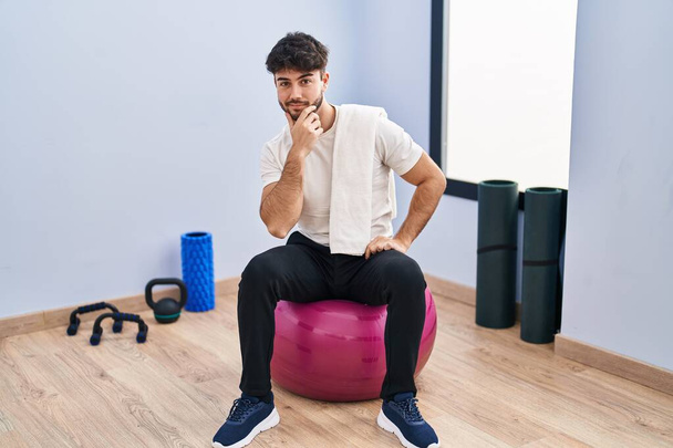 Hispanischer Mann mit Bart sitzt auf Pilatebällen im Yoga-Raum und blickt selbstbewusst in die Kamera, mit verschränkten Armen und erhobener Hand am Kinn. Positives Denken.  - Foto, Bild