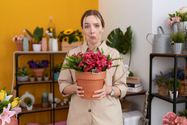 花屋で働く若い白人女性は、植物の鍋を保持し、苦痛のために落ち込んで心配し、怒って恐れて泣いています。悲しい表情.  - 写真・画像