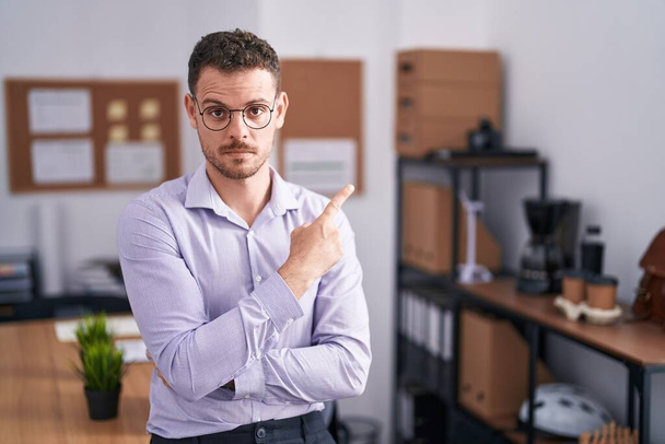 Joven hombre hispano en la oficina señalando con el dedo de la mano a un lado mostrando publicidad, cara seria y tranquila  - Foto, Imagen