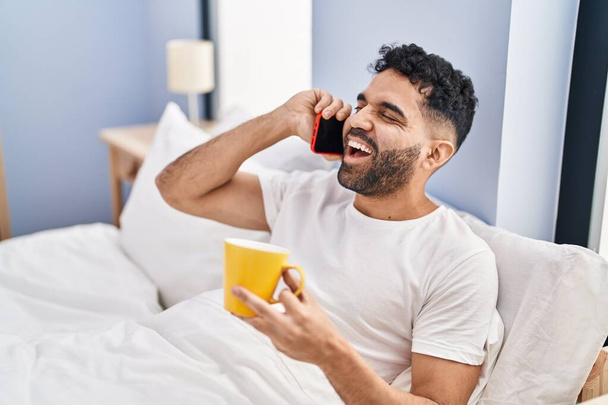 Fiatal spanyol férfi beszél okostelefon iszik kávét ül az ágyon a hálószobában - Fotó, kép
