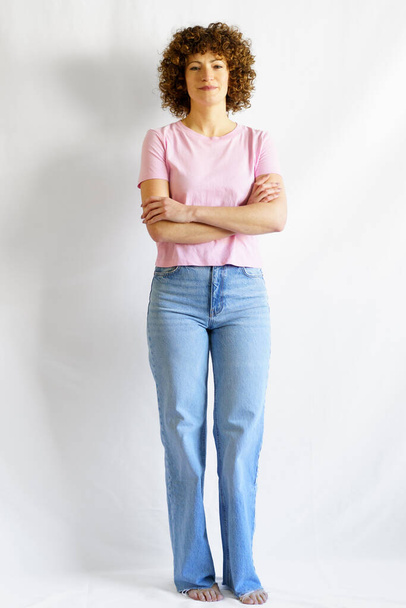 Full body of curly haired female in jeans and pink basic t-shirt leaning on white wall with hands crossed looking at camera - Foto, immagini