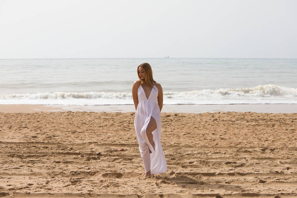 Nuori kaunis vaalea nainen valkoisessa mekossa kävelee hiekalla rannalla aurinkoisena päivänä. Nainen tekee erilaisia kehon ilmeitä. Taustalla sininen meri. - Valokuva, kuva