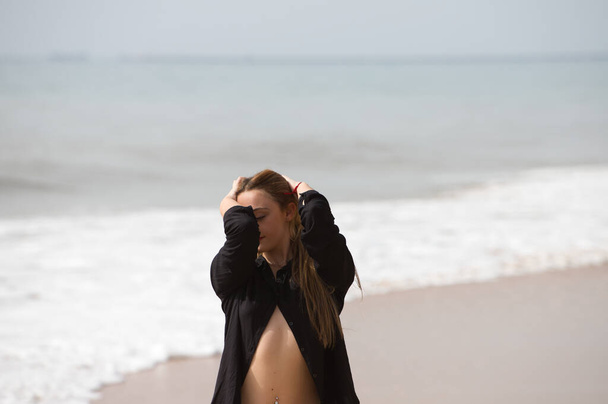 若くて美しいブロンドの女性は黒のシャツと黒のビキニを着たビーチの海岸に沿って歩き、異なる表情や姿勢を作ります。海と空を背景に. - 写真・画像