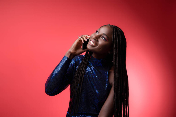 Smiling young black female in sequin dress talking on cellphone, happy millennial african american woman with braids hairstyle enjoying pleasant phone call while standing over red background - Photo, Image