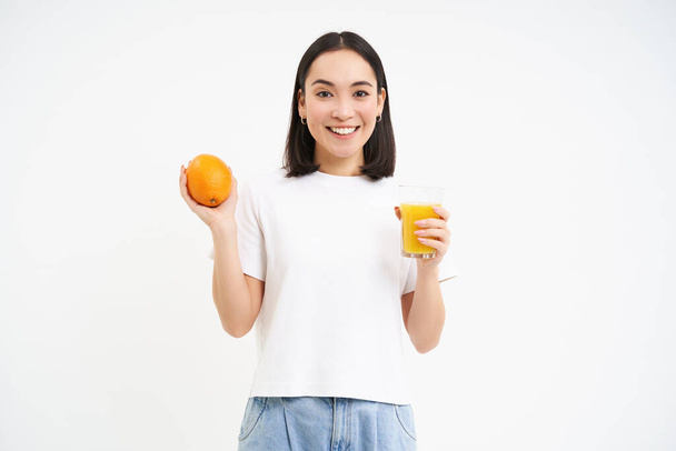 Uśmiechnięta Koreanka trzyma pomarańczowy fuit i sok, koncepcja dobrego samopoczucia i zdrowego odżywiania, białe tło. - Zdjęcie, obraz