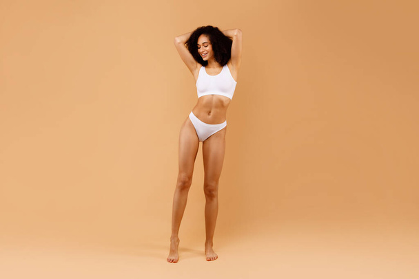 Full length shot van sensuele jonge zwarte dame poseren in ondergoed met handen achter haar hoofd over beige studio achtergrond, tonen perfecte lichaamsvorm, kopieer ruimte - Foto, afbeelding
