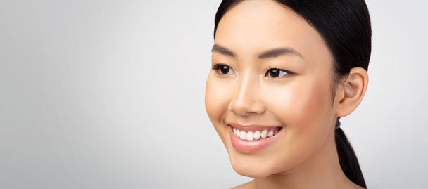Female Beauty. Closeup Portrait Of Happy Japanese Young Lady Smiling Looking Aside At Empty Space For Text, Advertising Products For Face Skin Care Over Light Gray Studio Background. Panorama - Zdjęcie, obraz
