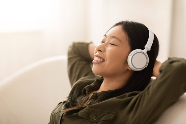 Closeup of pretty young asian woman relaxing on couch with hands behind head and closed eyes, using wireless stereo headphones at home, listening to music or audiobook, copy space - Foto, immagini