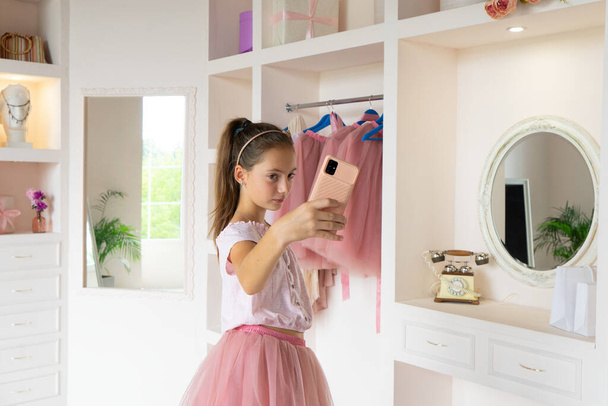 Rear view of tender teen girl in pink tutu-skirt and black high-heeled shoes with cell phone posing in front of mirror in wardrobe room, looking at own reflection and making selfie on smartphone - Fotó, kép