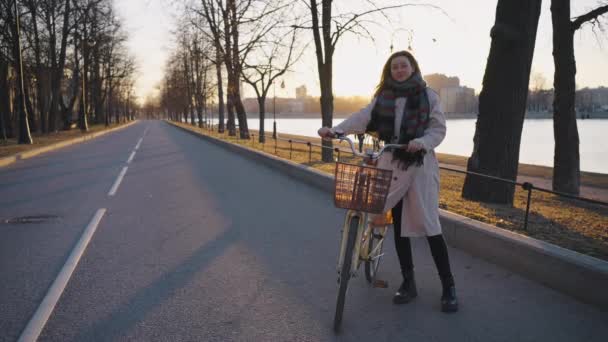 Mujer con estilo en una bufanda caliente y gabardina se levanta con una bicicleta de crucero con una cesta en una carretera vacía en un parque en el terraplén de la ciudad, un retrato al atardecer a principios de primavera, cámara lenta - Imágenes, Vídeo