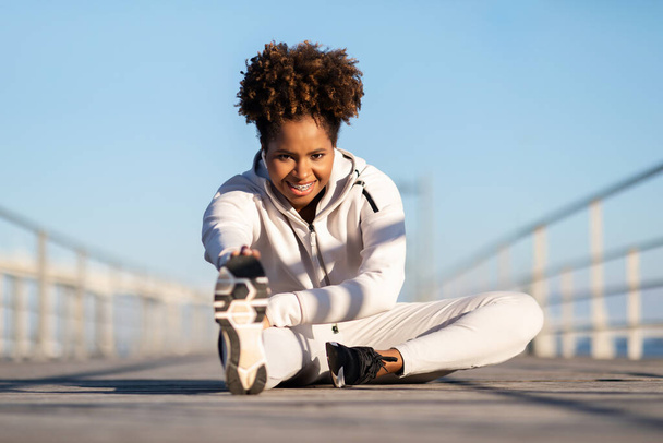 Афиши на улице. Улыбающаяся чёрная спортивная женщина разогревается перед тренировкой, молодая афроамериканская спортсменка в спортивной одежде растягивает мышцы ног во время упражнений на деревянном пирсе снаружи - Фото, изображение