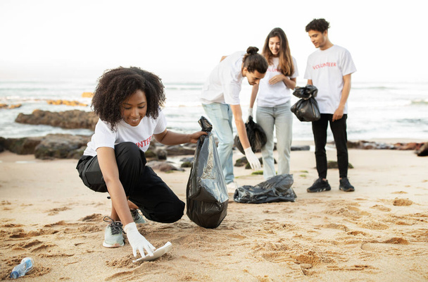 Ευτυχισμένοι νέοι πολυεθνικοί εθελοντές με μπλουζάκια με πακέτα σκουπιδιών καθαρίζουν τα σκουπίδια, πλαστικά μπουκάλια στη θάλασσα, υπαίθρια. Προστασία του περιβάλλοντος, προστασία του περιβάλλοντος - Φωτογραφία, εικόνα