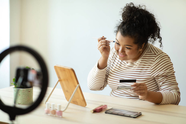 Krásný blog, blogovací koncept. Veselý pěkný kudrnatý mladá žena plus velikost v ležérní oblečení sedí za stolem před zrcadlem doma, nahrávání videa při aplikaci make-upu, pomocí blogger set - Fotografie, Obrázek