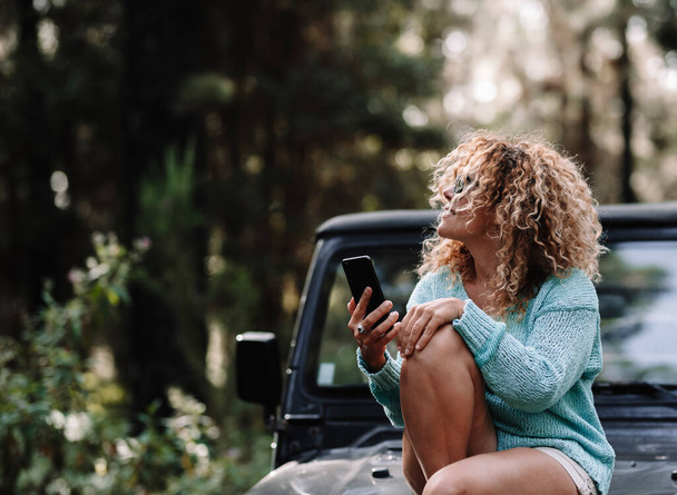  traveler woman enjoy outdoor vehicle leisure activity relaxing sitting on the front car and using connection technology with cellphone. Female people with nature forest and transport concept - Foto, imagen