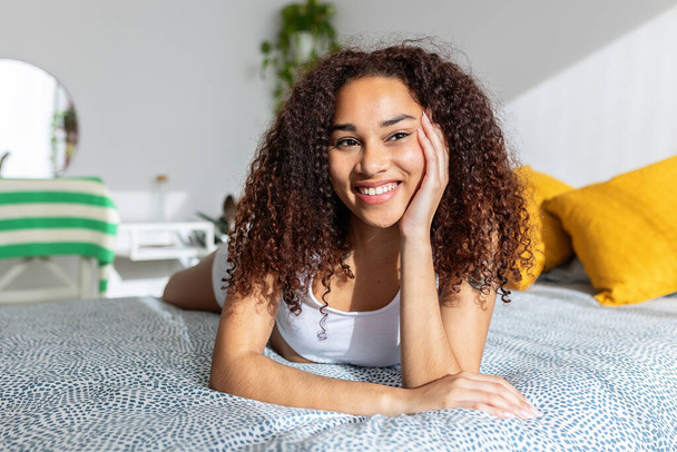 Usmívající se krásná mladá dospělá žena odpočívající v posteli. Portrét tisícileté latinské dívky s rukou na bradě dívá pryč, zatímco přemýšlí. - Fotografie, Obrázek