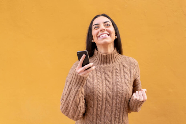 Jeune femme brune à l'extérieur en utilisant un téléphone mobile et en faisant un geste de victoire - Photo, image