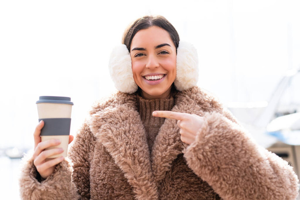 Mujer joven vistiendo manguitos de invierno y sosteniendo tomar café al aire libre y señalándolo - Foto, Imagen