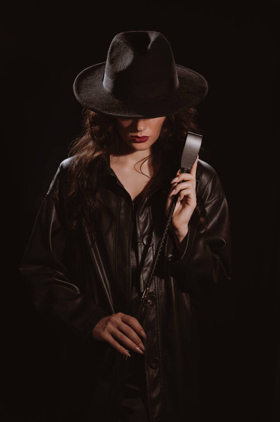 mujer en un impermeable de cuero y sombrero con un látigo flogger para el sexo BDSM con sumisión y dominación - Foto, Imagen