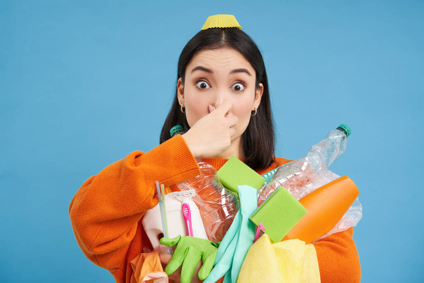 Portrait of woman shuts her nose from digust, holding stinky garbage, dirty plastic items and empty bottles with bad smell, standing over blue background. - Foto, afbeelding