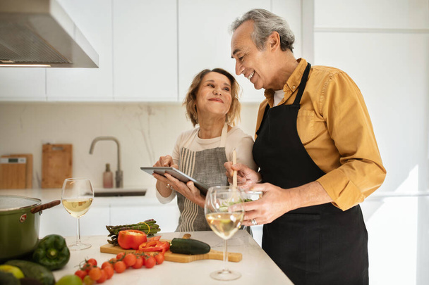 Senior caucasian couple preparing healthy dinner together, using digital tablet in cozy kitchen interior, free space. Healthy food at home, great offer, receipt and ad, relationships - Foto, Bild