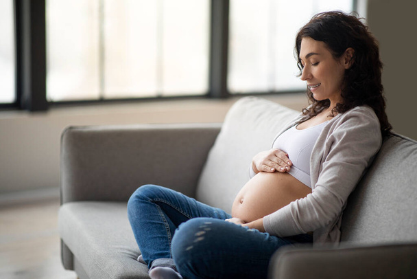 Готовимся к материнству. Молодая беременная нежно касается живота, расслабляясь дома на диване, красивая ожидающая мать ласкает живот и улыбается, наслаждаясь временем беременности - Фото, изображение