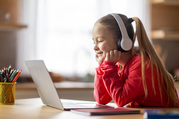 Hymyilevä pieni valkoihoinen tyttö kuulokkeissa kannettavan tietokoneen avulla kotona, söpö onnellinen esiteini naispuolinen lapsi opiskelee tietokoneella, ottaa online-oppitunnin tai luokan, nauttii kotiopetuksesta, kopioi tilaa - Valokuva, kuva