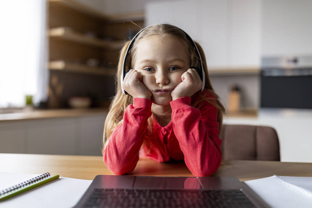 Langweiliger Inhalt. Pov-Aufnahme eines aufgebrachten kleinen Mädchens mit drahtlosen Kopfhörern, das zu Hause auf den Laptop-Bildschirm schaut, gelangweiltes frühgeborenes weibliches Kind, das am Schreibtisch sitzt und Computer benutzt, Standpunkt der Webcam - Foto, Bild