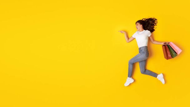 Wow Sale. Happy Woman Advertising Shopping Offer Posing With Shopper Bags And Pointing Finger At Free Space For Text On Yellow Studio Background. Fashion And Commerce. Panorama - Photo, image