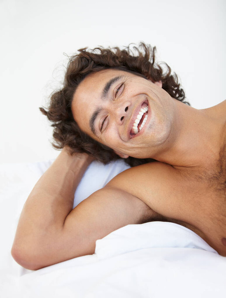 Face, laugh and a handsome man in bed in his home to relax or rest alone on a summer morning. Smile, wellness and lifestyle with a happy young male person relaxing in the bedroom of his house. - Foto, afbeelding