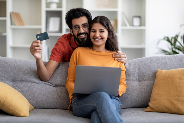 Концепция покупок онлайн. Счастливые индийские супруги с помощью ноутбука и кредитной карты дома, веселые молодые восточные семейные пары покупки в Интернете во время отдыха с компьютером в гостиной, скопировать пространство - Фото, изображение