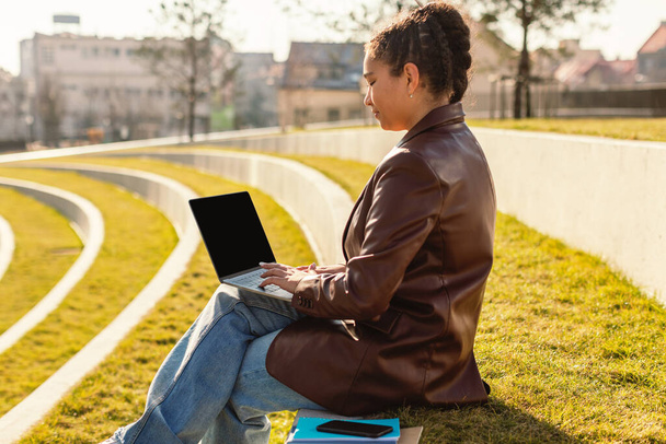 Female mixed race student using laptop computer with blank screen outdoors, sitting on lawn in park and looking at monitor, mockup for educational website - Photo, Image
