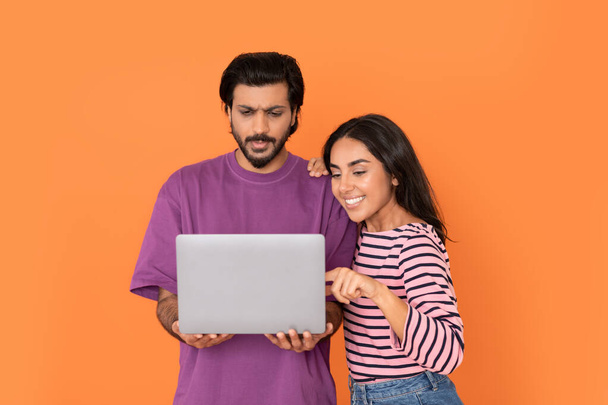 Beautiful cheerful loving young indian couple shocked handsome bearded man and pretty long-haired woman using pc laptop on orange studio background, shopping online, wife choosing new expensive dress - Photo, Image