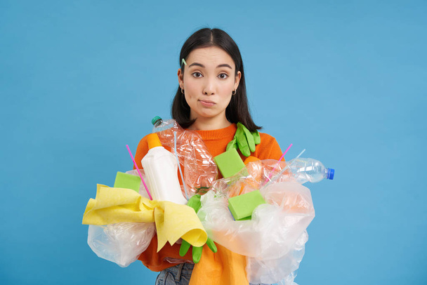 Young confused asian girl with different types of plastic, learns how to recycle, saving enviroment, sorting garbage, blue background. - Zdjęcie, obraz