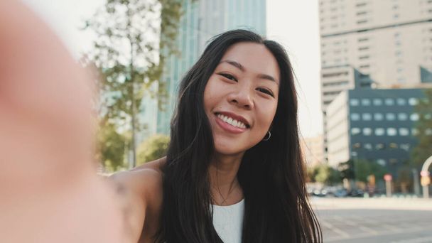 Close up, cute happy girl dressed in white top and jeans makes selfie on mobile phone while standing on modern city background - Fotoğraf, Görsel