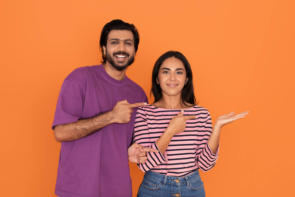Loving cheerful beautiful young indian couple in casual hindu man and woman pointing at free space above female hand, smiling at camera, isolated on orange studio background - Photo, Image