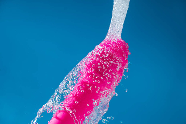 Pink vibrator under running water on a blue background. Sex toy hygiene concept - Foto, afbeelding