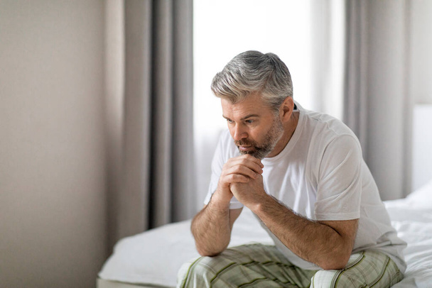 Hombre guapo de pelo gris de mediana edad deprimido que usa pijama sentado en la cama solo en casa, apoyado en sus manos, sufriendo de soledad o ansiedad, espacio para copiar. Permacrisis, crisis de mediana edad - Foto, Imagen