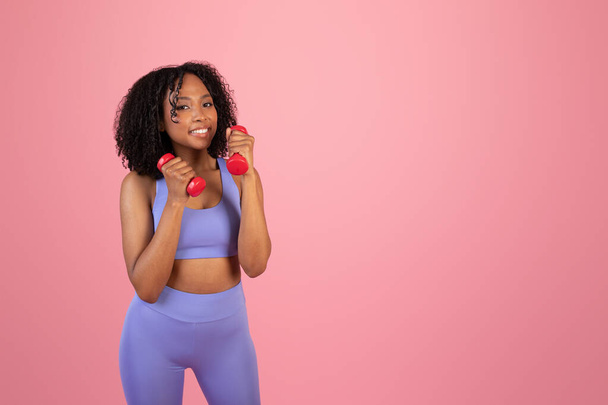 Glad millennial african american lady in sportswear doing exercises with hand dumbbells, enjoy workout isolated on pink background, studio. Sports recommendation, ad and offer, fitness with coach - Photo, Image