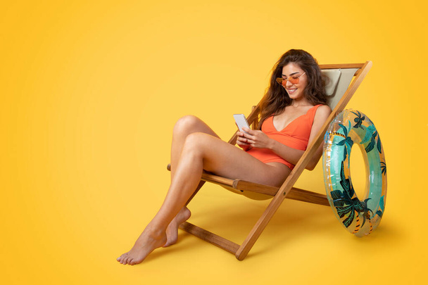 Usmívající se tisíciletá Asiatka v plavkách, sluneční brýle s nafukovacím kroužkem sedí na lehátku, užívají si volného času, chatují po telefonu izolovaném na oranžovém pozadí. Léto s gadgetem, odpočinek a relax - Fotografie, Obrázek