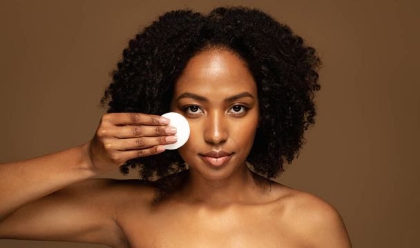 Sensual smiling pretty naked young black woman using cotton pad, hydrating face skin with toner, erasing makeup with micellar water, brown studio background, copy space, closeup - Photo, Image
