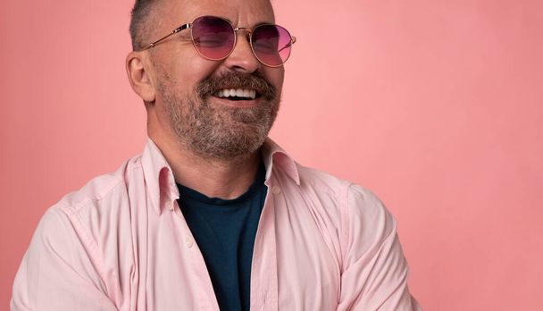 Portrait of a successful handsome smiling mature man in rose glasses dressed in pastel pink shirt isolated on pink background. handsome bristle man face with big white smile - Φωτογραφία, εικόνα